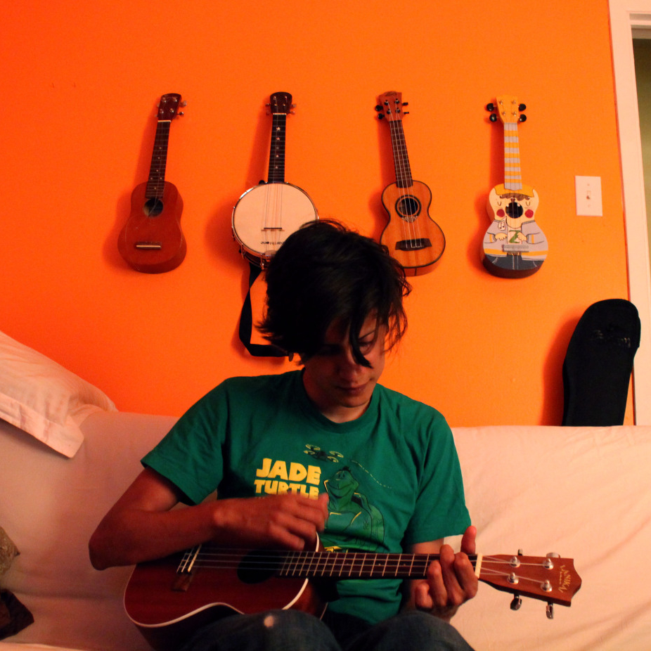 an image of Juan Camilo playing the ukulele, while sitting on a white couch, in a room with an orange wall